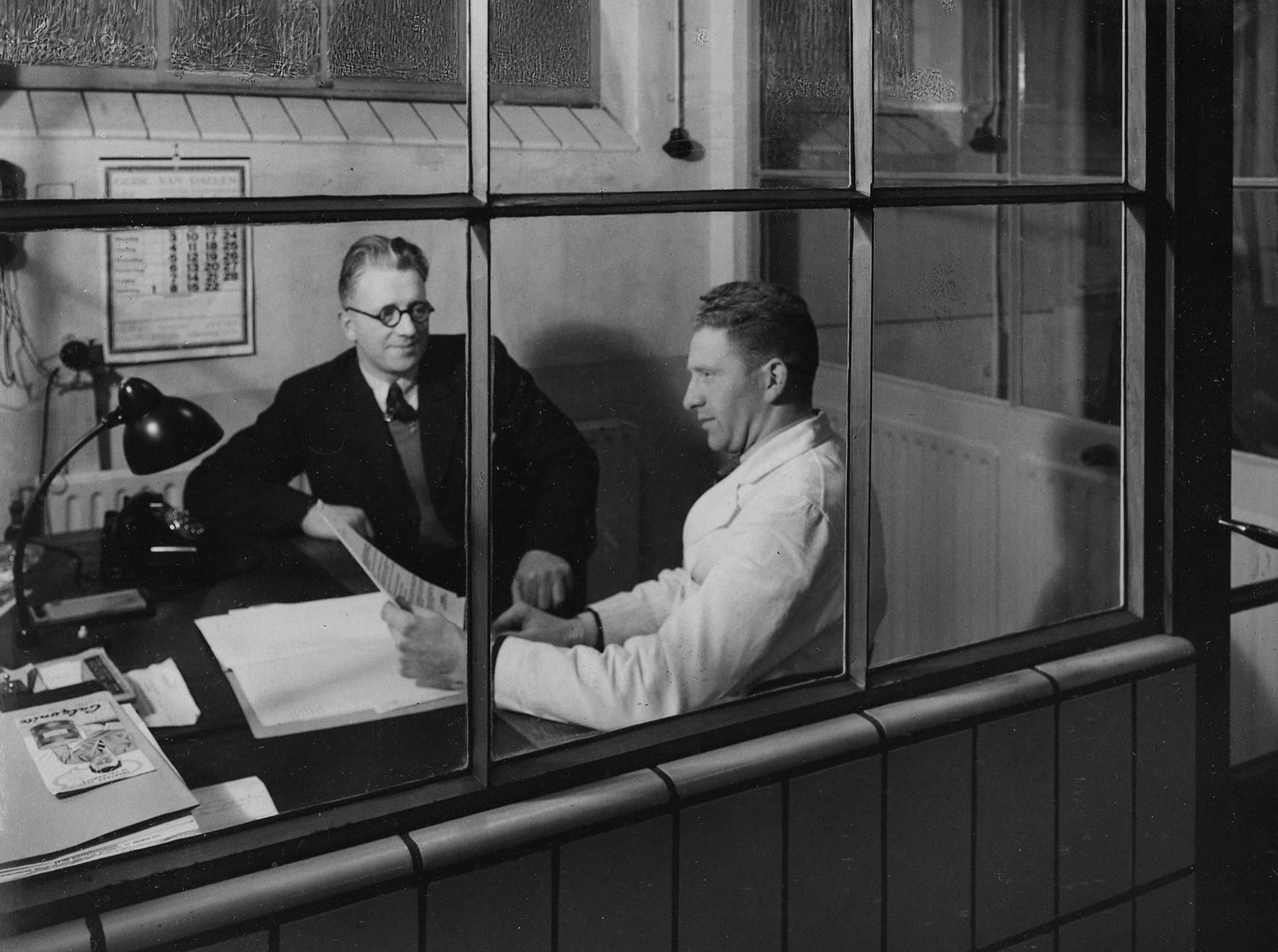 Foto de arquivo em preto e branco de dois homens conversando em um escritório, separados por uma divisória de vidro